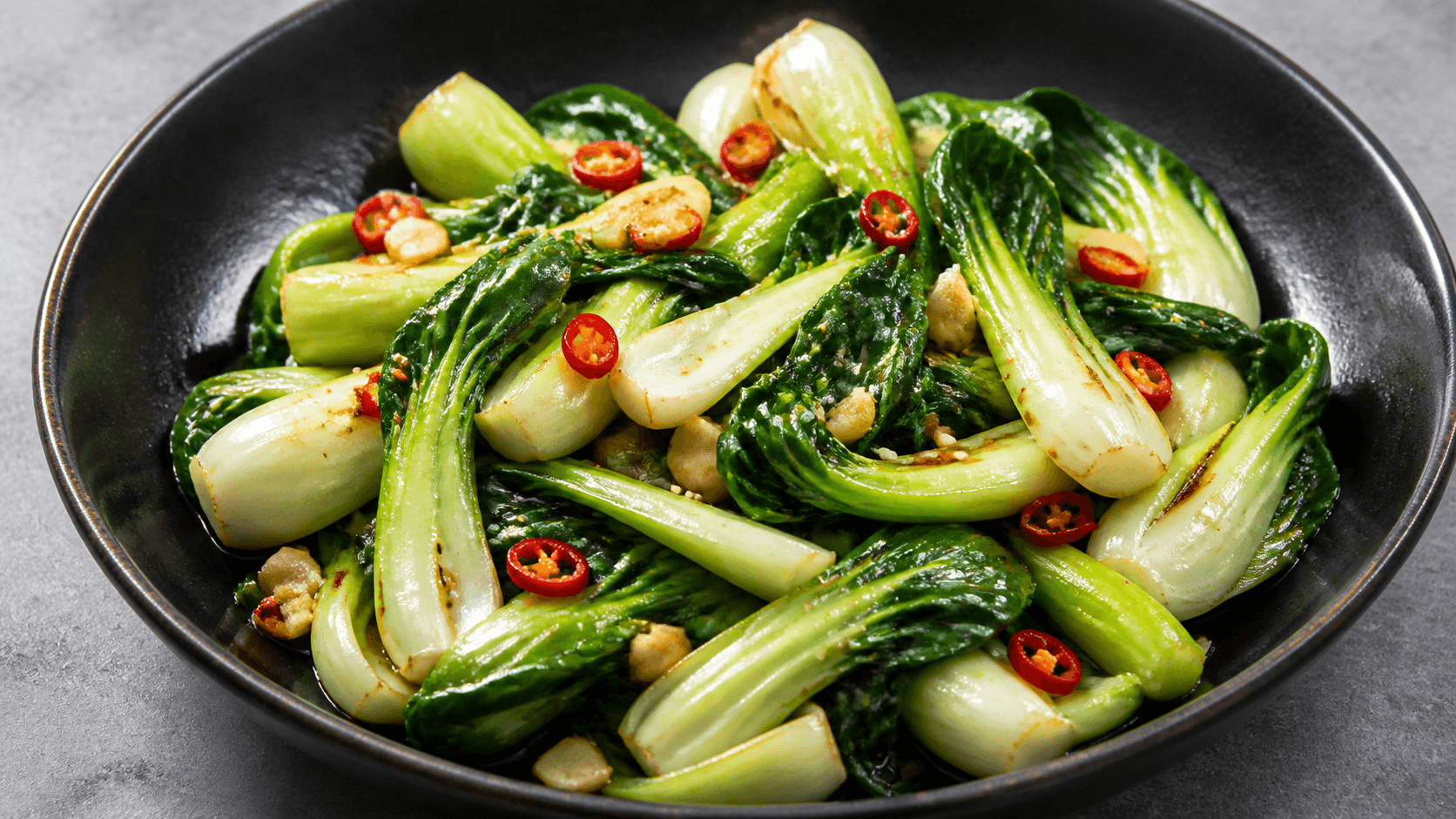 Pak choi wok i mørk skål med blanke grønne blade, hvide stilke, chili og sesam.