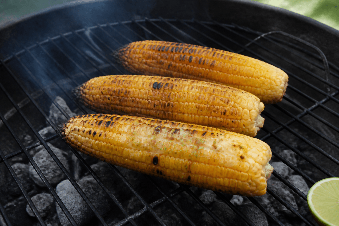 Tre grillede majskolber med ujævne mørke grillmærker og chili-lime-smør på en kulgrill.
