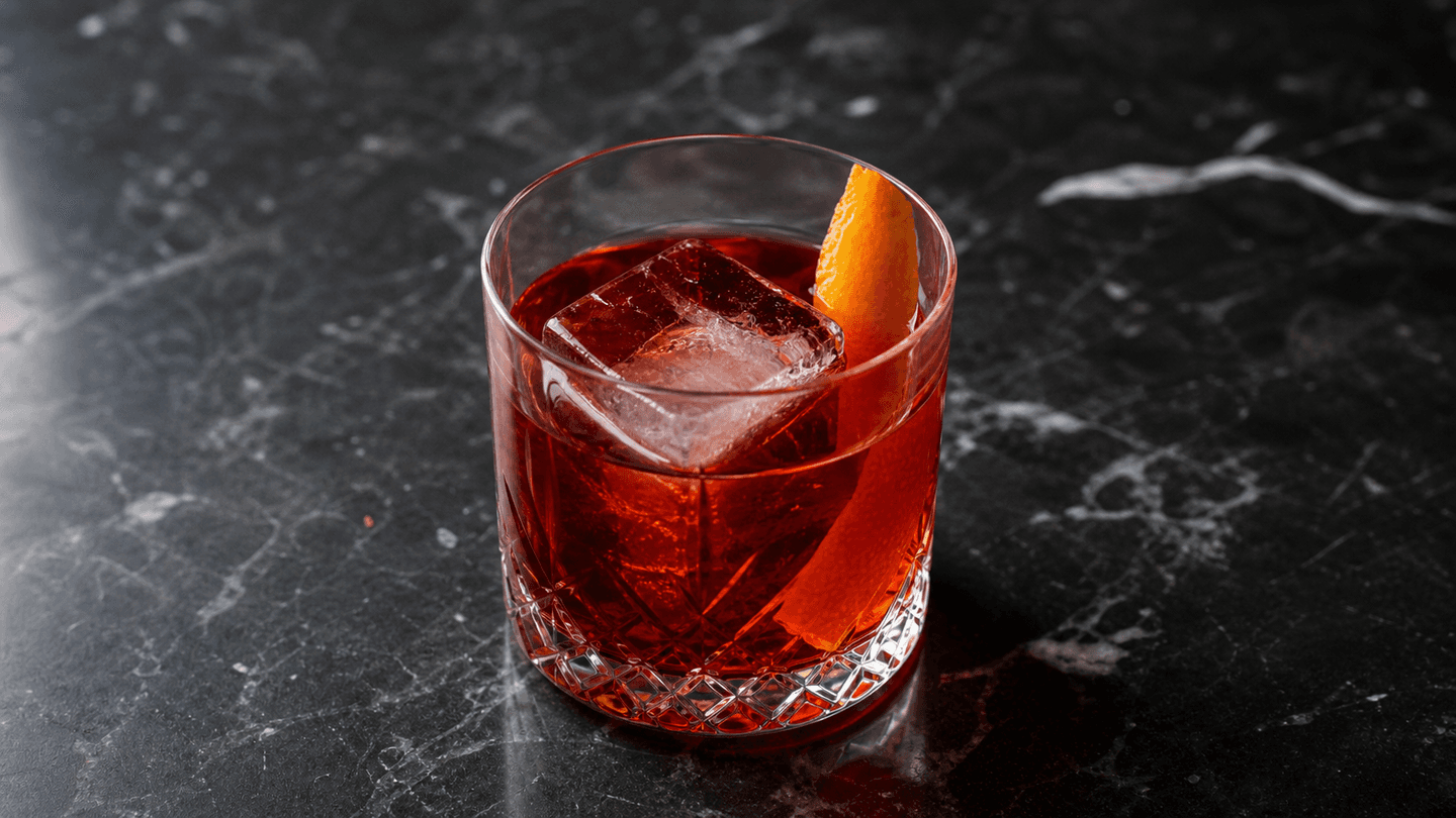 Negroni i lavt glas med stor isterning, rubinrød drink og appelsinskal.