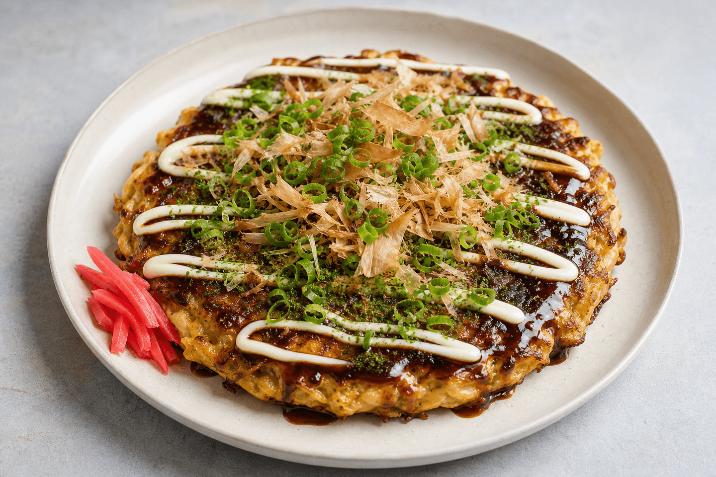 Okonomiyaki på tallerken med brun sauce, mayo-striber, grønne urter, bonito og syltet ingefær.