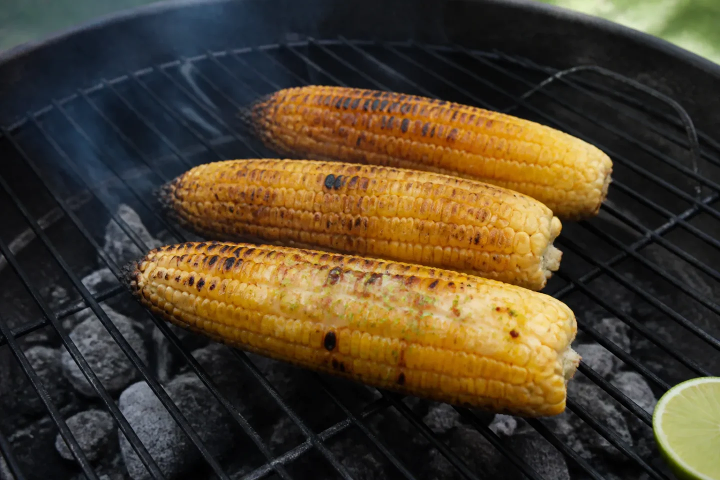 Tre grillede majskolber med ujævne mørke grillmærker og chili-lime-smør på en kulgrill.