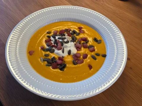 Cremet orange hokkaidosuppe i hvid tallerken med en skefuld creme fraiche, græskarkerner og sprøde bacontern.